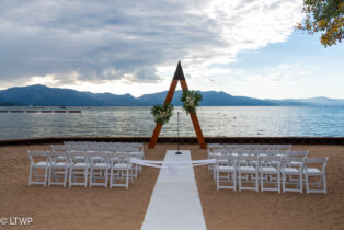 Outdoor wedding setup on a sandy beach with white chairs, a wooden arch, and a scenic view of a lake and mountains in the background.