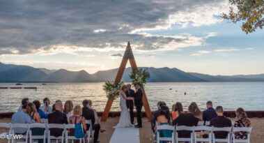 A couple exchanges vows at a beachfront wedding ceremony under a triangular arch, with guests seated and mountains in the background at sunset.