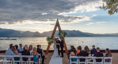 A couple exchanges vows at a beachfront wedding ceremony under a triangular arch, with guests seated and mountains in the background at sunset.
