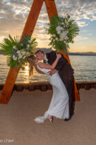 A couple in wedding attire kisses passionately under a triangular floral arch by a lakeside at sunset.