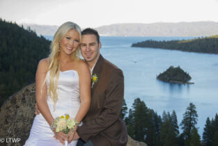 A newlywed couple smiling, seated with a scenic view of a lake and island in the background. the bride holds a bouquet.