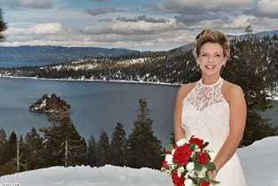 A bride in a white gown holding a bouquet of red flowers, standing in snow with a forested lake and mountains in the background.