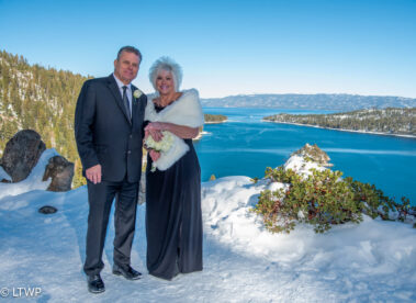 A couple in formal attire, the man in a suit and the woman in a gown with a fur stole, stands on a snow-covered overlook by a lake under a clear blue sky.