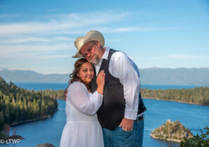 A couple in formal wear with cowboy hats embrace by a lake with a forested island in the background.