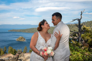 A couple in wedding attire affectionately facing each other at a scenic lakeside location with forested hills and blue skies in the background.