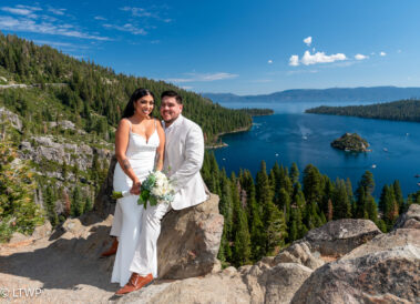 Newlywed couple posing on a rocky overlook with a panoramic view of a lake and forested islands under a clear blue sky.