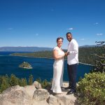 A couple standing on top of a mountain near the water.