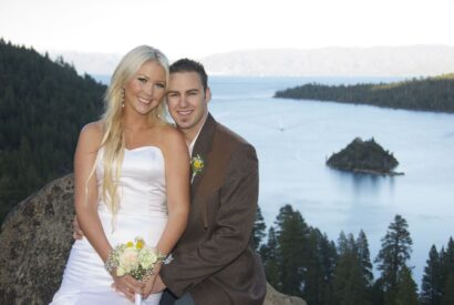 A newlywed couple smiling and posing outdoors with a scenic lake and a small island in the background.