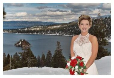 A bride holding a bouquet of red flowers, smiling, with a snow-covered landscape and lake in the background.