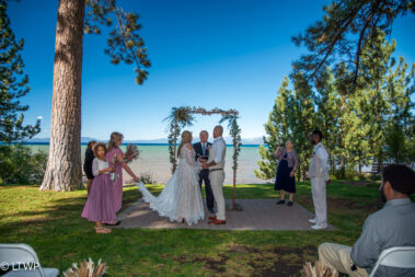 Outdoor wedding ceremony by a lake with a couple exchanging vows under an arch, attended by a small group of guests.
