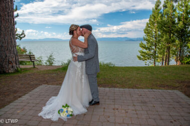 A bride and groom kissing by a lakeside, surrounded by trees and a clear sky, with distant mountains in the backdrop.
