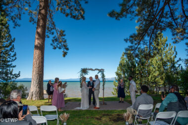 A wedding ceremony by a lake with guests seated under tall trees, a couple exchanging vows under a floral arch.
