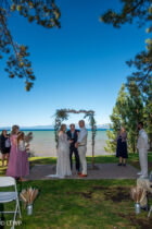 A wedding ceremony taking place outdoors by a lake, with a couple standing under an arch, surrounded by guests and trees.