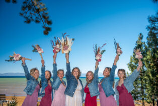 Seven women in pink dresses and denim jackets joyfully hold bouquets in the air, standing outdoors with a clear blue sky and lake in the background.