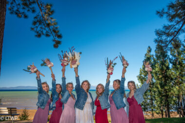 Bride and bridesmaids in denim jackets holding bouquets, posing with arms raised against a lake and tree backdrop.