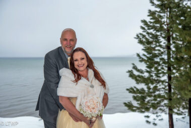 A couple in wedding attire posing outdoors with a lake and snow-covered landscape in the background.