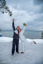 A couple celebrating in snow near a lake, the man in a black jacket and the woman in a white outfit, raising one leg and holding flowers.