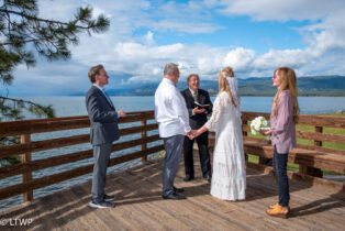 A wedding ceremony on a wooden deck overlooking a lake, with two couples and an officiant standing under a clear blue sky.