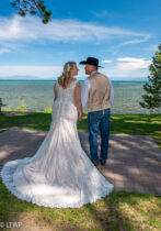 A bride in a lace gown and a groom in a cowboy hat and jeans stand hand in hand, looking at a lake surrounded by trees.