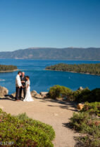 A bride and groom holding hands on a scenic lakeside overlook with clear blue sky and lush greenery.