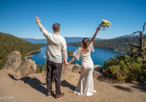 A newlywed couple holds hands, facing a lake, with the bride raising a bouquet in celebration, under a clear blue sky.