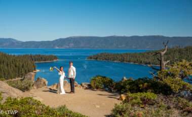 A couple in wedding attire stands on a mountain overlooking a panoramic view of a clear blue lake and surrounding forest.
