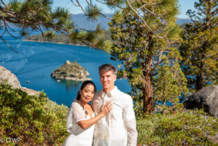 A couple in wedding attire stands by a lake with an island in the background, framed by pine branches, smiling at the camera.