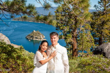 A couple in wedding attire stands by a lake with an island in the background, framed by pine branches, smiling at the camera.