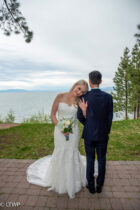 A bride and groom stand together, facing a scenic lake with trees,