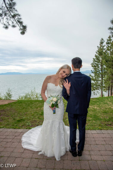 A bride and groom stand together, facing a scenic lake with trees,