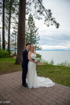A couple in wedding attire embracing near a lake with trees in the background.