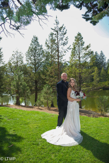 A bride and groom smiling and standing close together on grass, surrounded by pine trees,