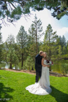 A bride and groom embrace outdoors on a grassy area beside a lake,