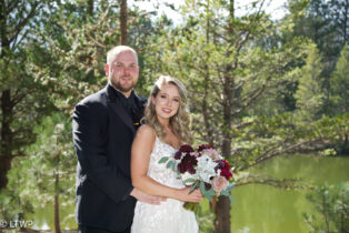 A bride and groom smiling outdoors with a lake and pine trees in the background.
