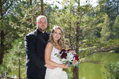 A bride and groom smiling outdoors with a lake and pine trees in the background.