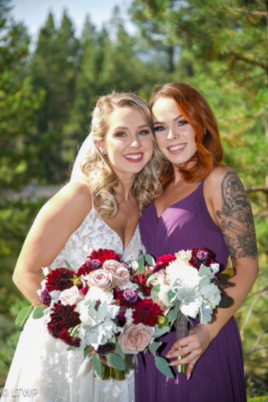 Two women smiling in a forest, holding a bouquet of flowers together.