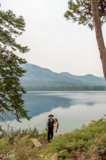 Bride and groom standing by a lake under a large tree, with mountains in the background shrouded by a hazy sky.