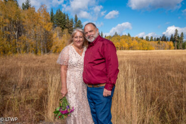 A happy couple poses in a field with autumn trees in the background, the woman in a floral dress and the man in a red shirt and jeans, holding hands.